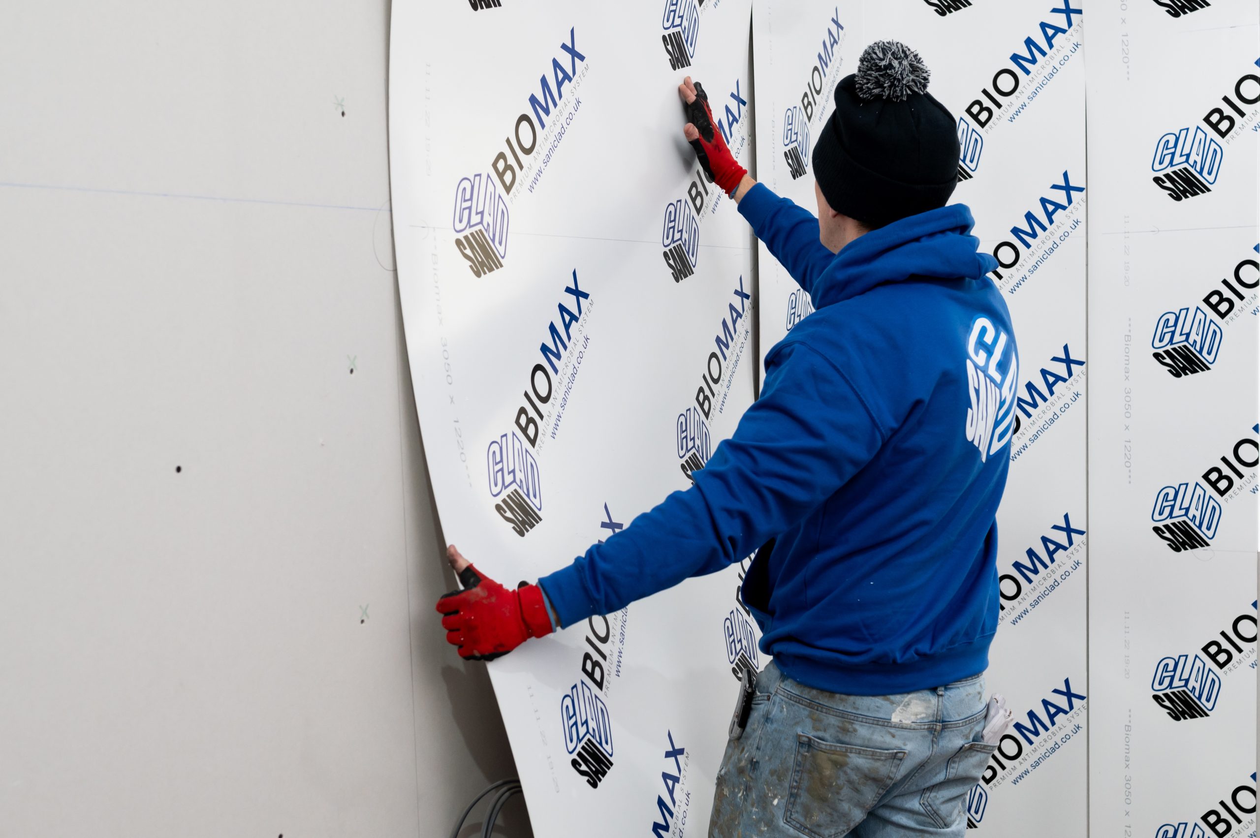 Worker in a blue hoodie and red gloves installing a large, flexible SANICLAD BIOMAX cladding panel onto a wall.